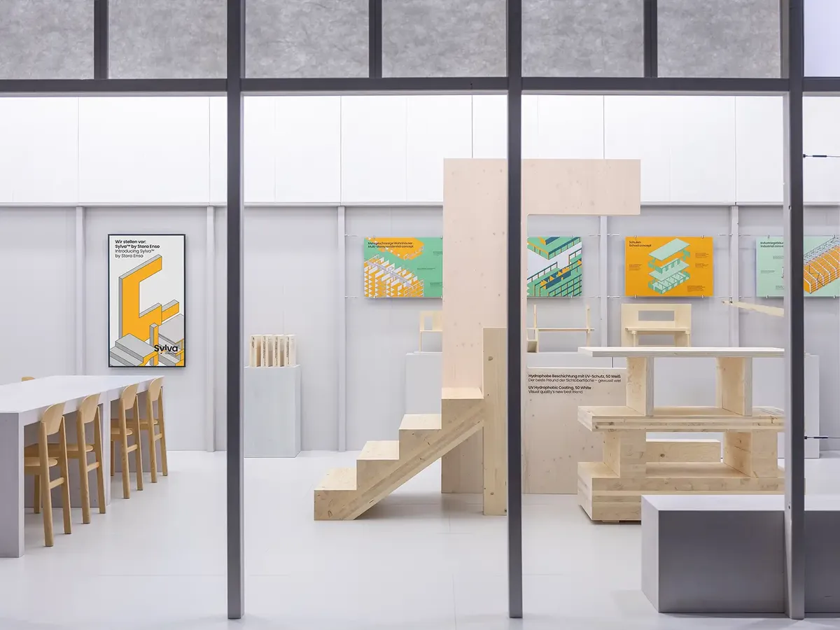 Stora Enso stand at BAU 2025 with a minimalist design in white and wood, showing technical illustrations on the wall and timber constructions as exhibits, complemented by a bar area with light wooden chairs.